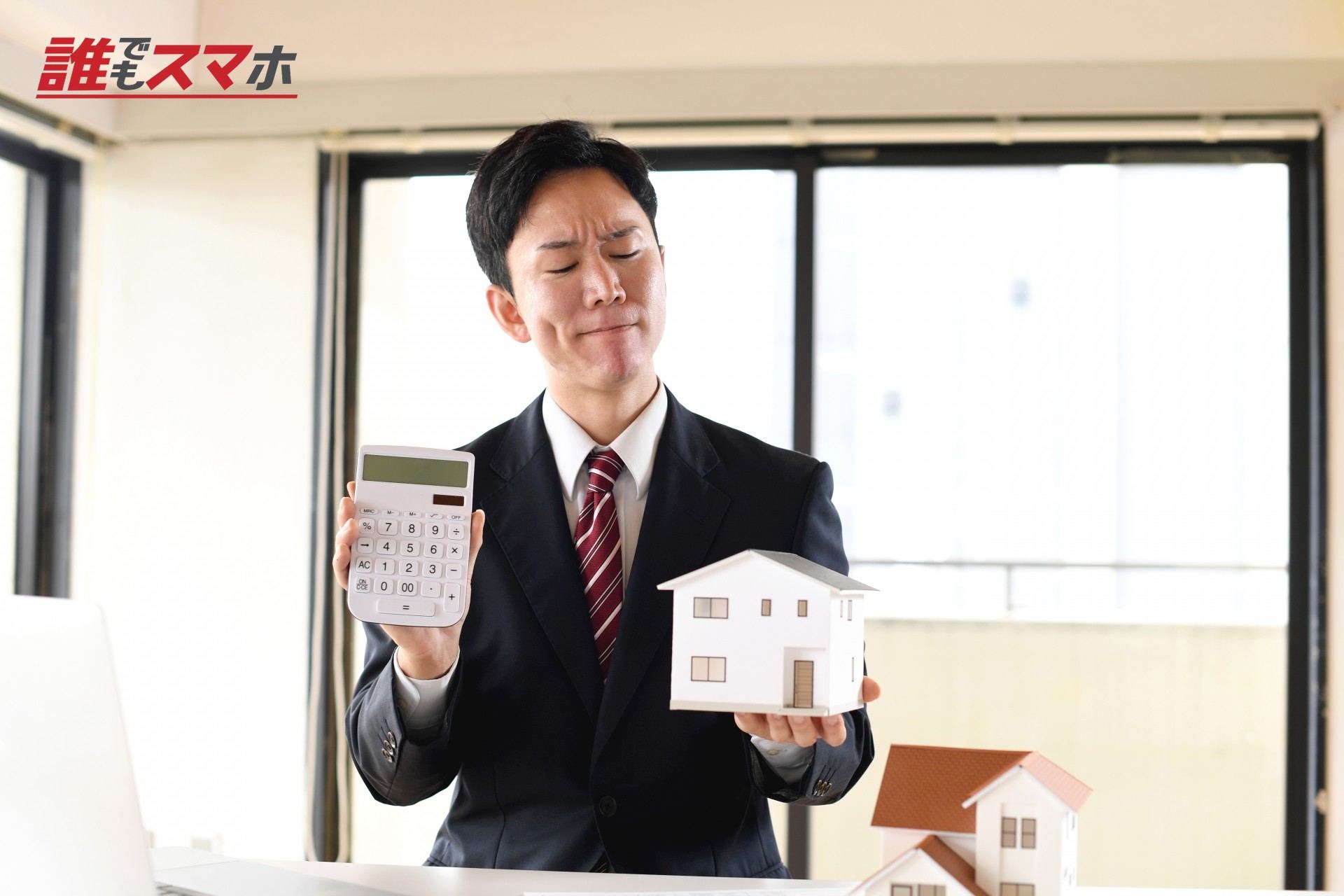 住宅模型と電卓を持ち悩むスーツ姿の男性